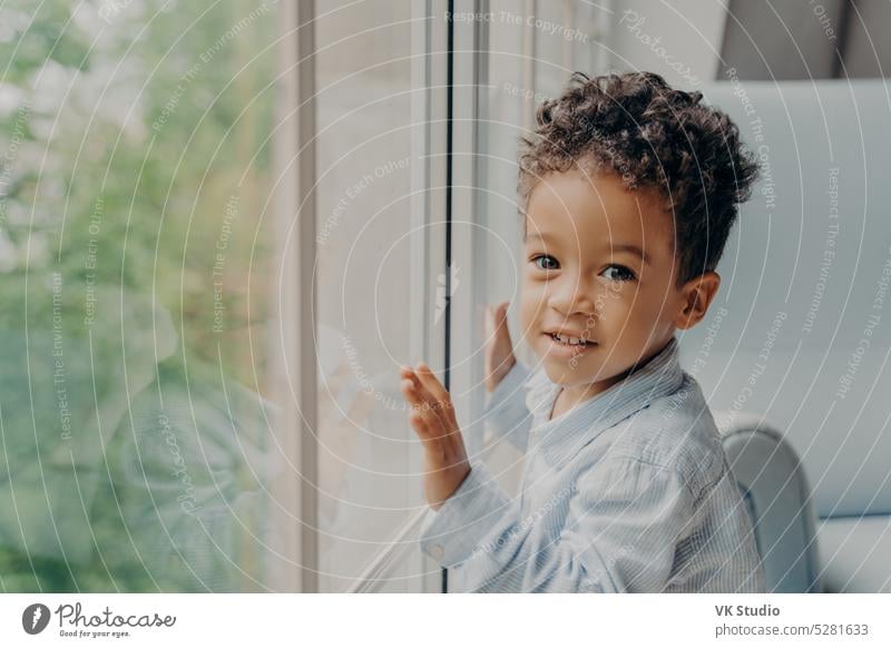 Cute mixed race little curly haired boy in light blue colored shirt next to large window kid looking son home wait parent toddler touching afro american