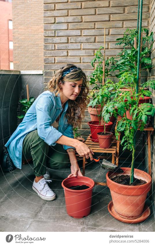 Woman using shovel to fill with soil a pot of plant in urban garden on terrace woman gardener female throwing potting plants rooftop transplanting gardening