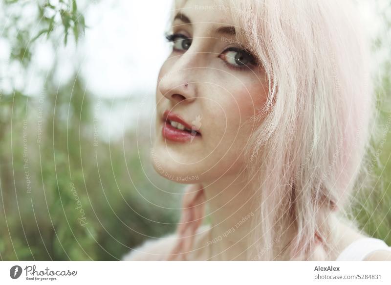 Young woman with pink blonde hair looks sideways at camera and stands in front of lake with trees and bushes on shore Woman portrait pretty Slim long hairs