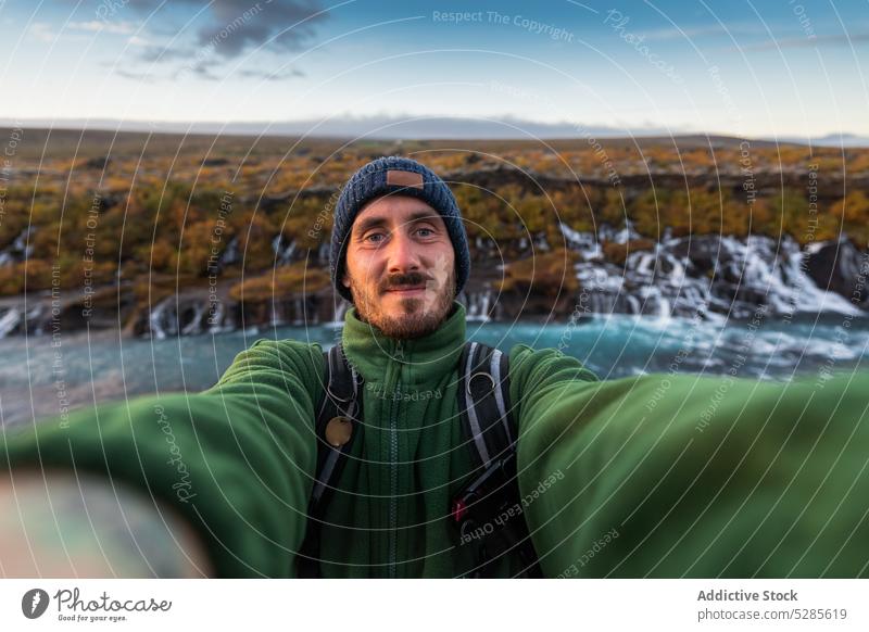 Positive traveler taking selfie near waterfall man hiker tourist self portrait cascade autumn mountain nature iceland male smile hat warm clothes outerwear