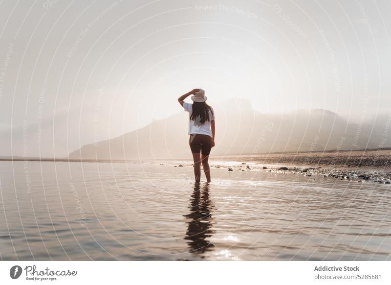 Anonymous woman walking in sea water near mountain tourist shore beach summer vacation nature coast spain asturias hat ocean sun seashore female sky seaside