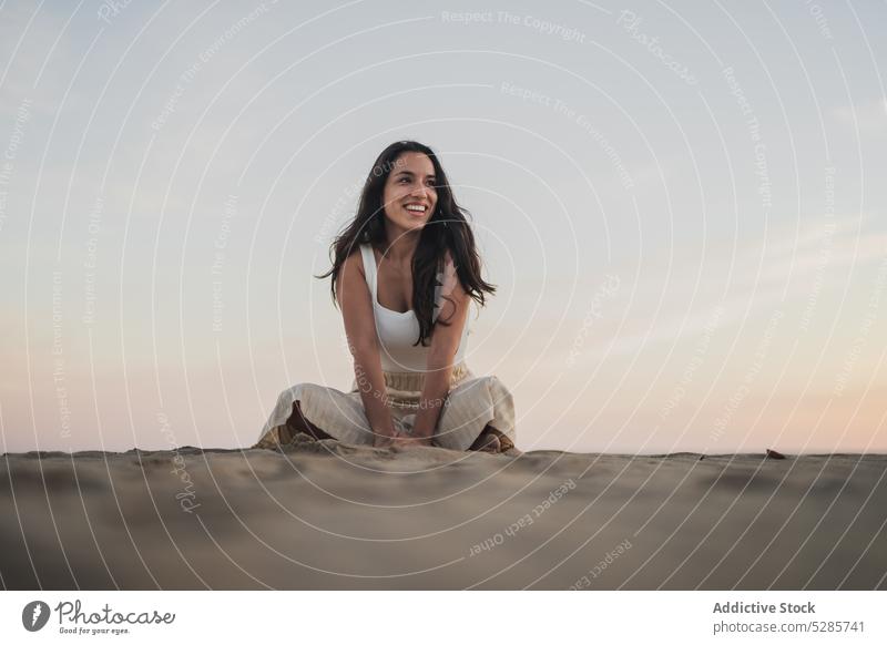 Carefree young ethnic woman sitting on beach at sundown smile sand holiday sunset legs crossed chill tourist happy positive summer vacation female hispanic