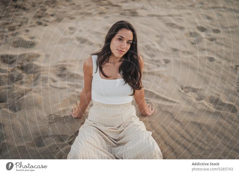 Carefree young ethnic woman sitting on beach at sundown smile sand holiday sunset legs crossed chill tourist happy positive summer vacation female hispanic