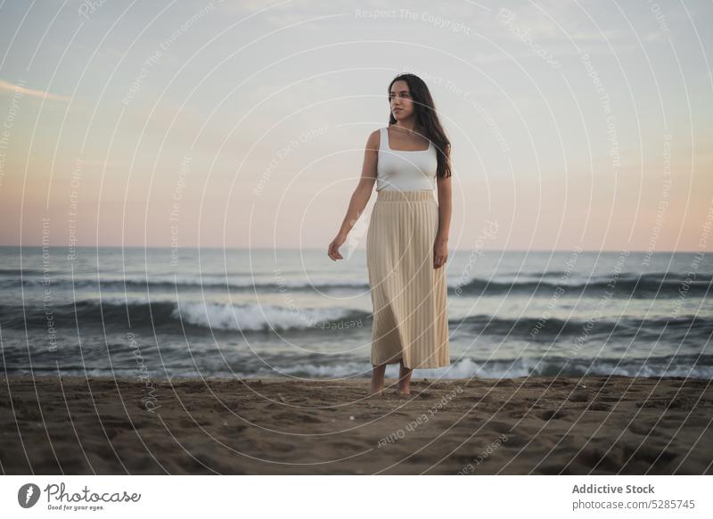 Cheerful young ethnic woman standing on sandy seashore during summer vacation walk beach enjoy holiday sunset traveler nature female hispanic barefoot long hair