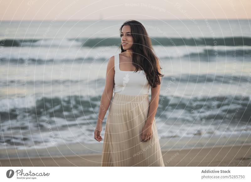 Cheerful young ethnic woman standing on sandy seashore during summer vacation walk beach enjoy holiday sunset traveler nature female hispanic barefoot long hair