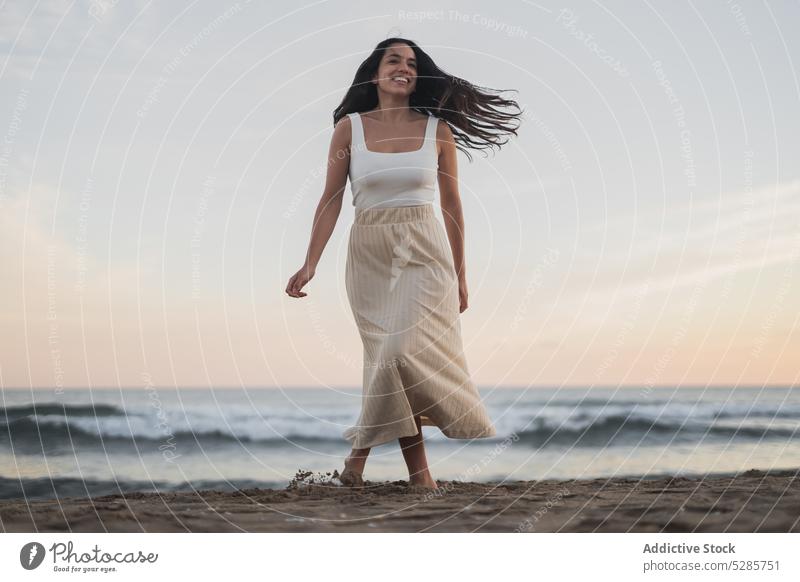 Cheerful young ethnic woman walking on sandy seashore during summer vacation beach smile enjoy happy holiday sunset traveler nature female hispanic barefoot