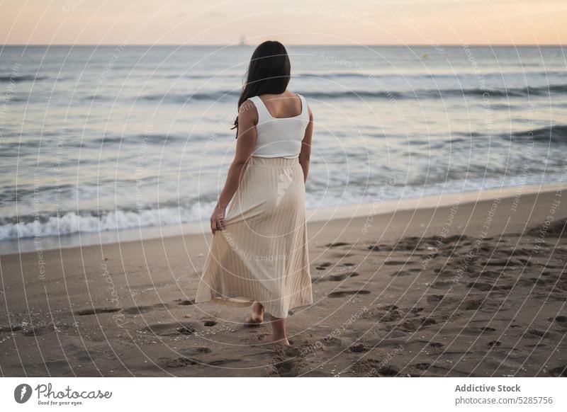 Anonymous young ethnic woman standing on sandy seashore during summer vacation walk beach enjoy happy holiday sunset traveler nature female barefoot long hair