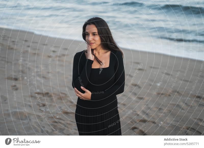 Delighted young ethnic female tourist standing on beach and smiling woman self assured vacation holiday feminine sea rest smile touch cheek portrait style