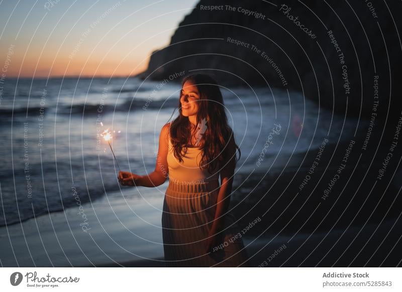 Smiling woman with sparkler on seashore sunset burn evening smile happy celebrate cheerful bengal female coast light festive casual ocean joy dusk twilight