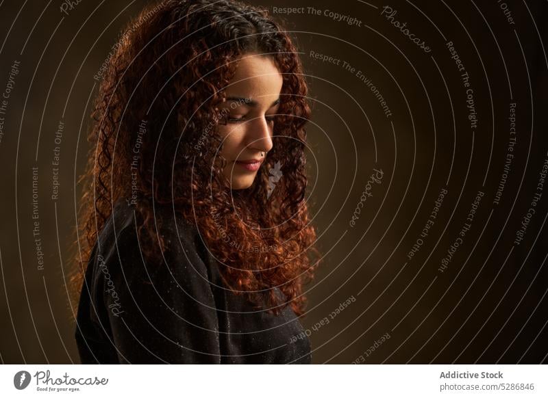 Charming woman standing in dark room thoughtful pensive calm serious tranquil appearance dreamy melancholy charming female style young curly hair brunette