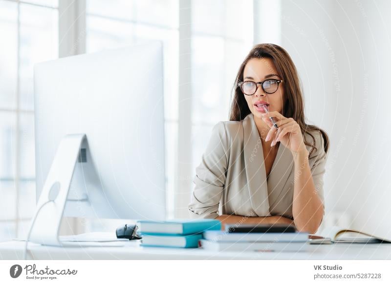 Contemplative female entrepreneur keeps pen in mouth, focused in monitor of computer, thinks on development of new strategy, wears elegant clothes and spectacles, poses against office interior