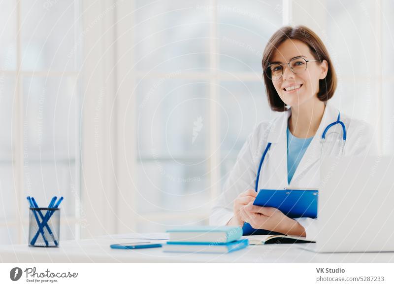 Female doctor writes prescription on special form, works in private clinic, wears white medical gown, ready for seeing patients, poses at workplace. Smiling physician or medical worker holds clipboard