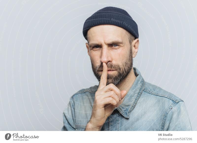 Serious pleasant looking bearded male with mysterious expression keeps fore finger on lips, asks to keep personal information confidential, wears stylish hat and denim shirt, isolated on grey wall