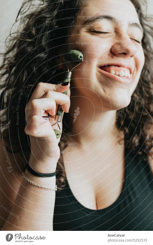 Smiling young woman using facial roller for skin care. Beauty healthcare habits with face massage. Getting ready and daily routine for glowing face. Closeup portrait in studio.