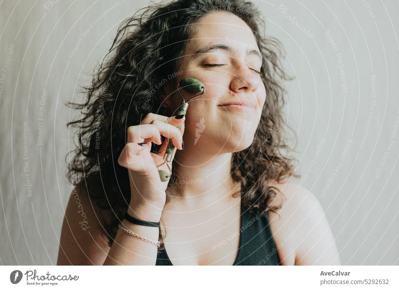 Smiling young woman using facial roller for skin care. Beauty healthcare habits with face massage. Getting ready and daily routine for glowing face. Closeup portrait in studio.