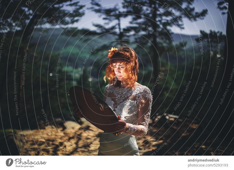 beautiful young red-haired bride in the forest with a floral wreath on her head and mirror . woman in long white dress outdoors on summer day. wedding day woods