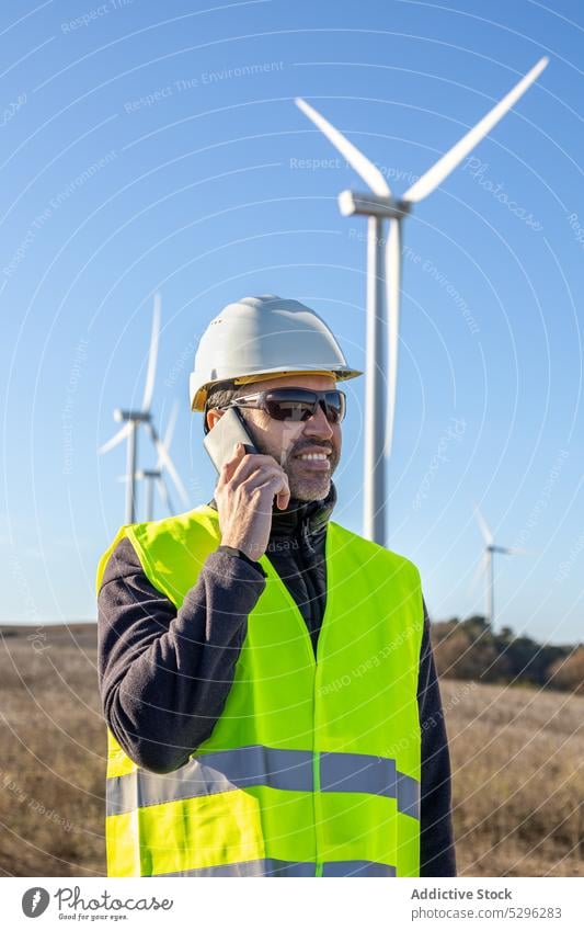 Engineer in protective helmet speaking on smartphone against wind turbines man engineer countryside roadway worker using phone call eco alternative energy