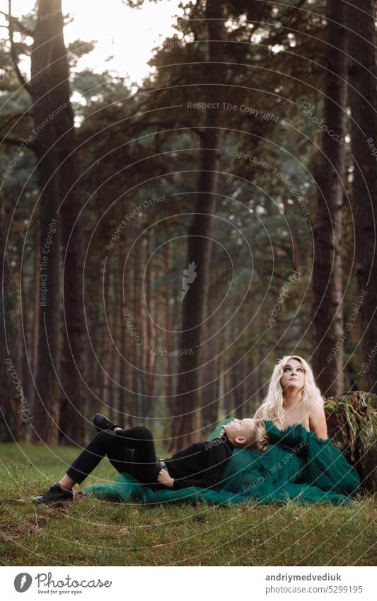 A beautiful blonde young woman in a long green dress and a diadem on her head with stylish young son in the forest. girl and boy sitting near the old stump. Solar glare. Fantasy. fairy tale