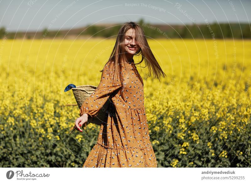 beautiful young woman walks in a field of yellow rapeseed. Girl brunette long hair fly in wind dressed in dress and straw hat. summer holiday concept female