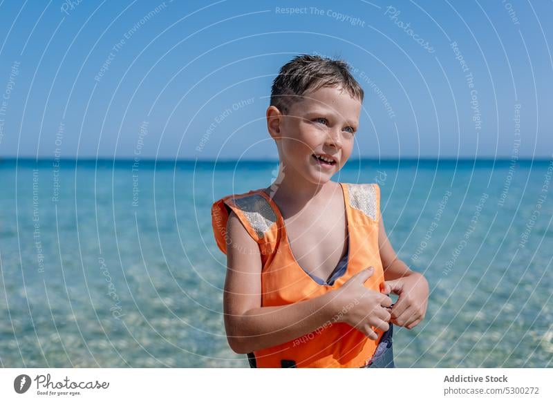 Smiling boy in swim safety jacket while standing near sea happy smile beach summer seashore child cheerful kid water joy glad coast childhood ocean positive