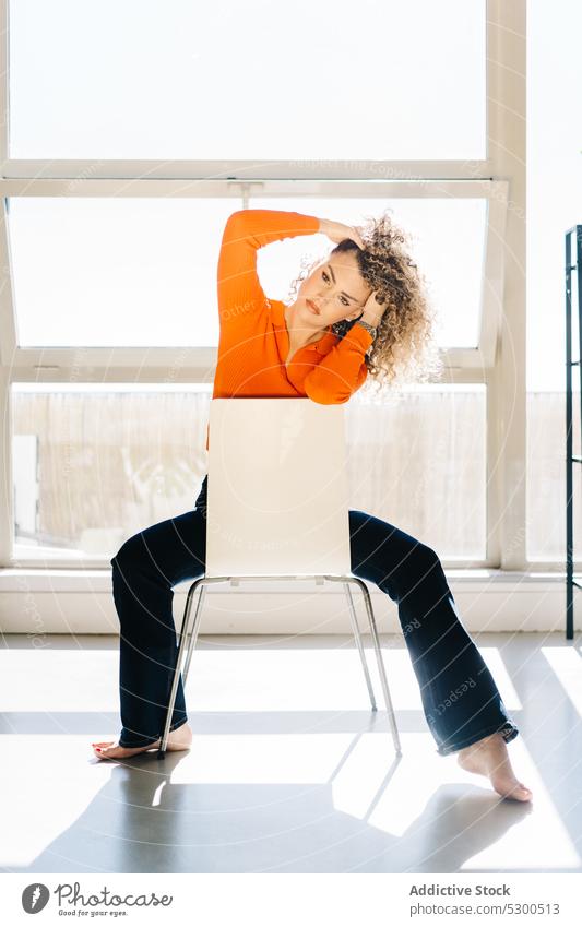 Carefree woman touching hair on chair touch hair confident rest barefoot style studio appearance trendy window female casual calm young relax charismatic