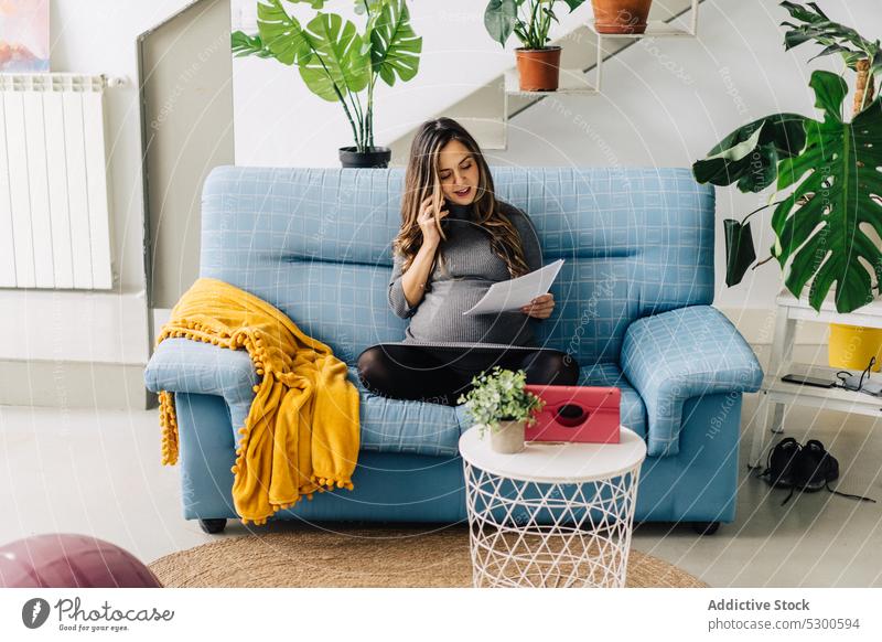 Smiling pregnant woman with documents having phone conversation using smartphone phone call apartment talk sofa smile female young work busy gadget communicate