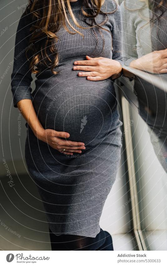 Anonymous pregnant woman standing near window caressing belly love touch belly maternity leave anticipate tranquil mother await female young stomach abdomen