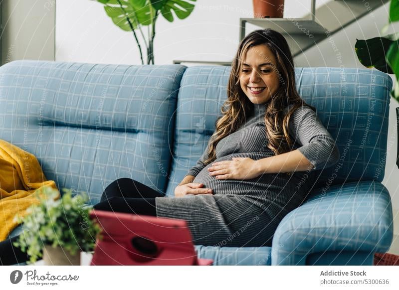 Smiling pregnant woman watching film on tablet movie maternity leave leisure free time sofa at home smile female young connection mother checkered wireless