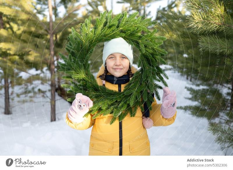 Cute preteen girl with wreath in snowy forest winter nature warm clothes smile coniferous hat cold woods season outerwear happy woodland wintertime positive