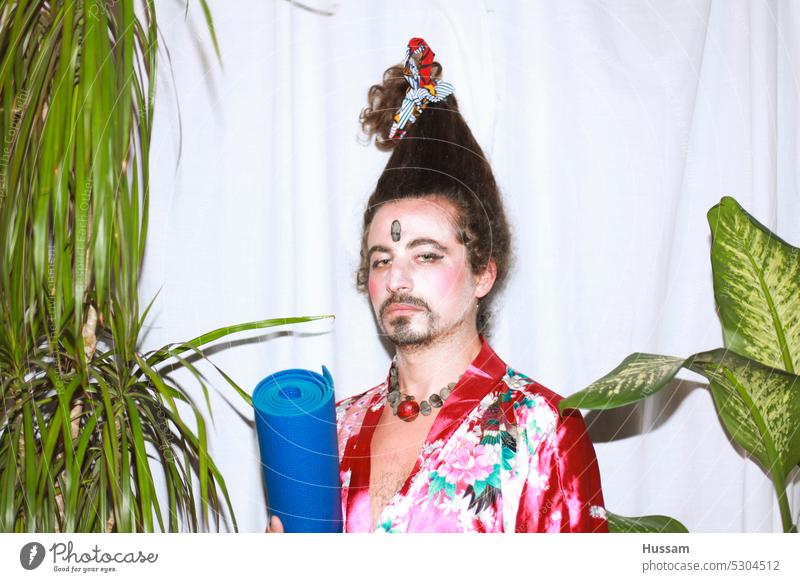 it is a photo concept about a man holding a yoga mat wearing a flowery dress and his hairy styled in a very funny way. His makeup  looks like a geisha with an ironic look.