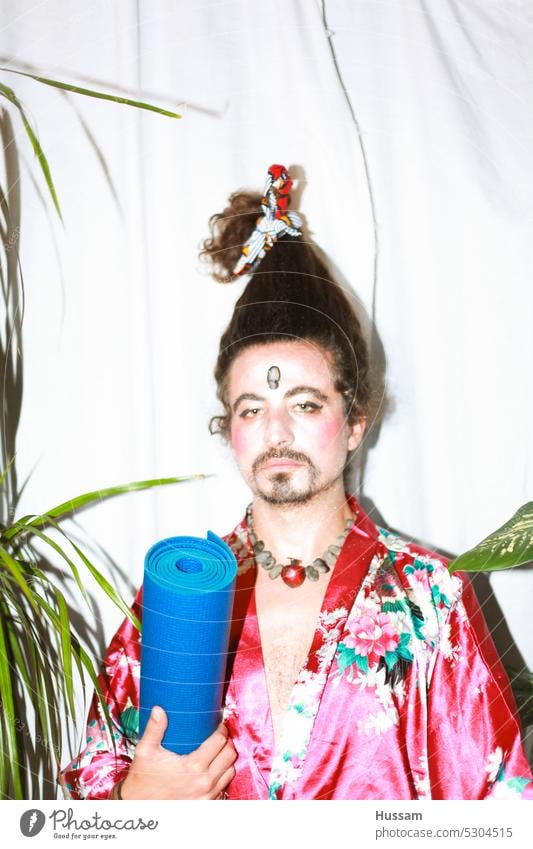 it is a photo concept about a man holding a yoga mat wearing a flowery dress and his hairy styled in a very funny way. His makeup  looks like a geisha with an ironic look.