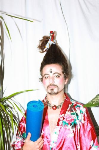 it is a photo concept about a man holding a yoga mat wearing a flowery dress and his hairy styled in a very funny way. His makeup  looks like a geisha with an ironic look.