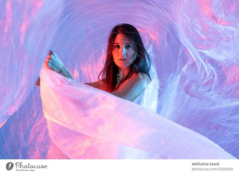 Woman looking at camera through plastic film woman cellophane neon light polyethylene pollute danger environment female disaster ecology ecosystem studio shot