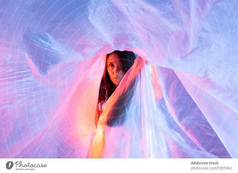 Woman looking at camera through plastic film woman cellophane neon light polyethylene pollute danger environment female disaster ecology ecosystem studio shot