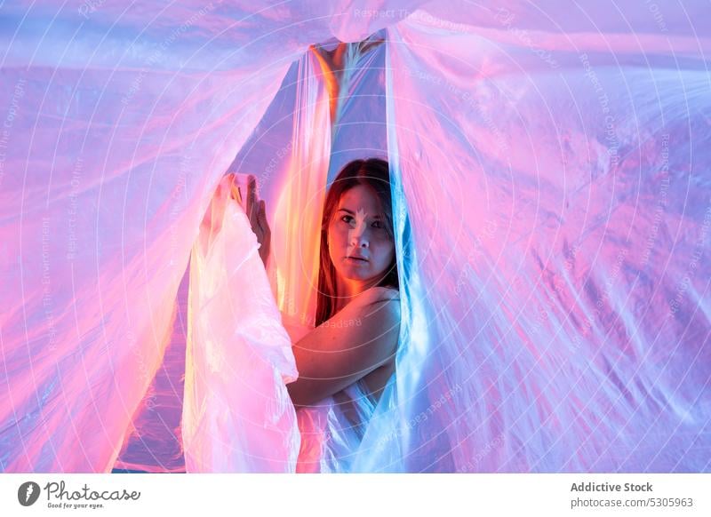 Woman looking at camera through plastic film woman cellophane neon light polyethylene pollute danger environment female disaster ecology ecosystem studio shot