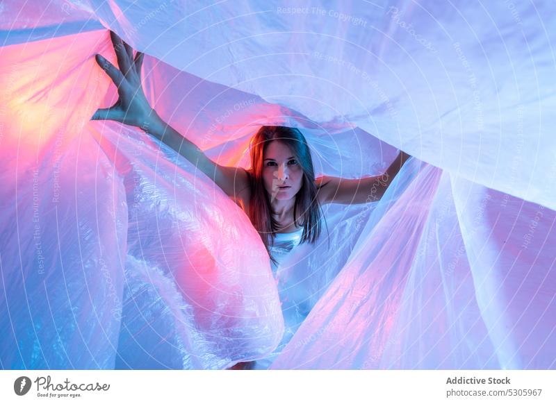 Woman looking at camera through plastic film woman cellophane neon light polyethylene pollute danger environment female disaster ecology ecosystem studio shot