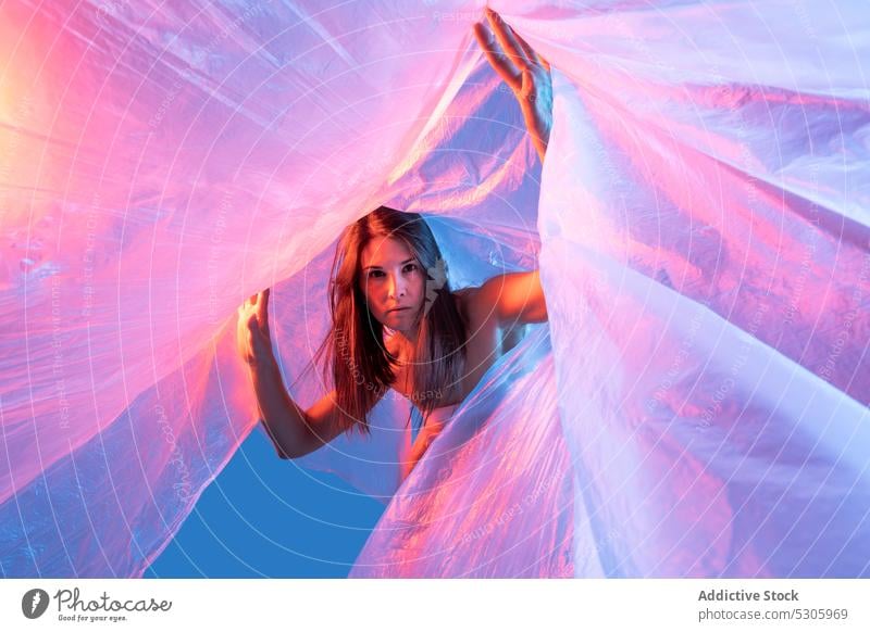 Woman looking at camera through plastic film woman cellophane neon light polyethylene pollute danger environment female disaster ecology ecosystem studio shot