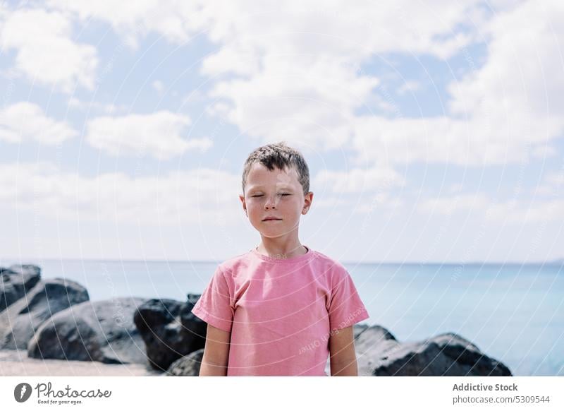 Cute boy standing on rocky seashore in daylight child summer coast thoughtful beach calm preteen blue sky childhood water seaside stone ocean kid cloudy