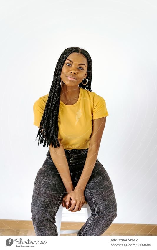 Smiling black woman sitting on chair against white wall smile positive trendy braid content appearance style confident female young casual ethnic calm