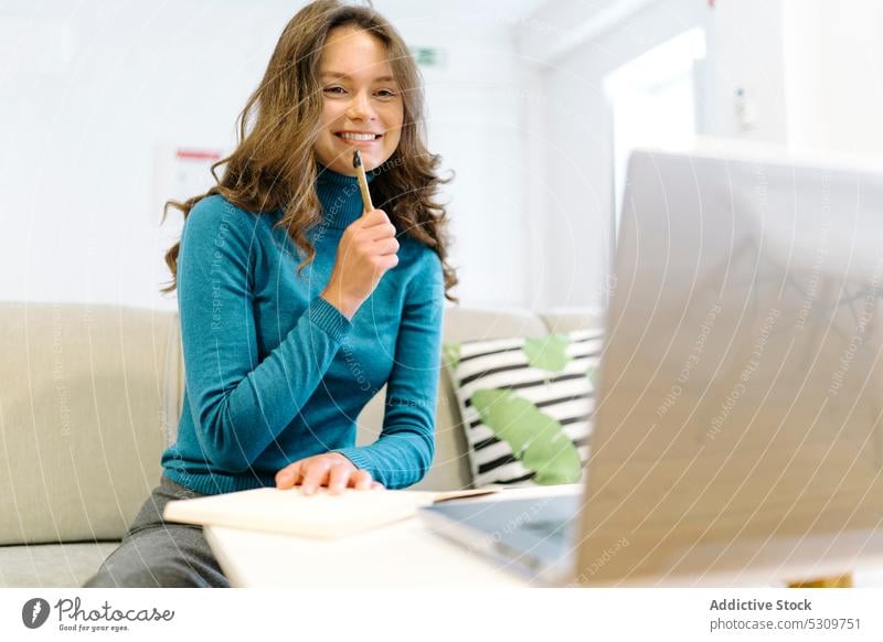 Thoughtful woman with pen taking notes in notebook near laptop on couch freelance pensive take note think project planner work remote female write young