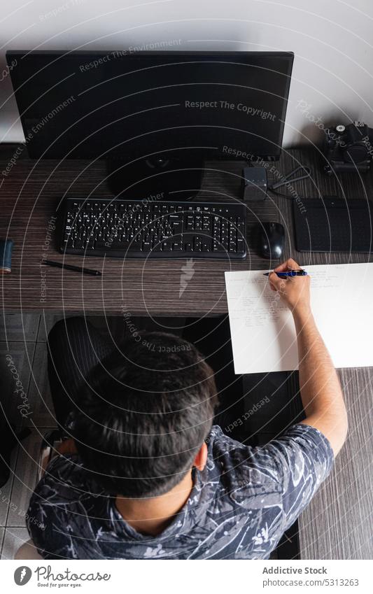 Man writing notes near computer man freelance work write take note paper workplace job workspace table device gadget male wooden photographer photo camera idea