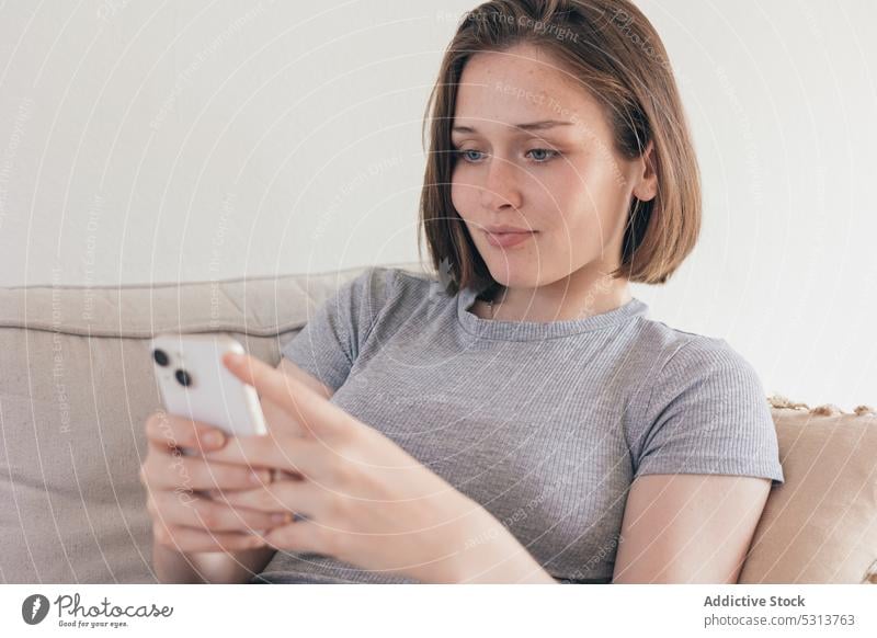 Content woman messaging via smartphone while sitting on sofa using home browsing domestic rest chill gadget couch device female living room mobile internet