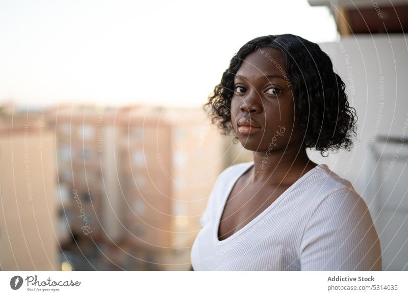 Calm black woman looking at camera against cityscape appearance balcony confident gaze portrait serious curly hair urban personality young female street calm