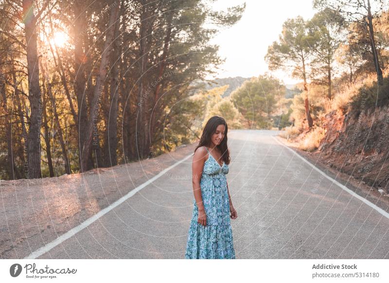 Carefree woman stand along road in forest traveler asphalt summer sunset city walk tree vacation female zaragoza admire spain dress street trip journey evening