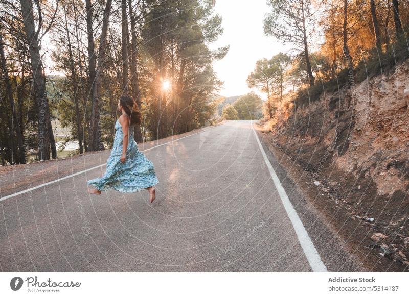 Carefree woman jumping along road in forest traveler asphalt summer sunset city tree vacation female zaragoza spain dress street trip journey evening barefoot