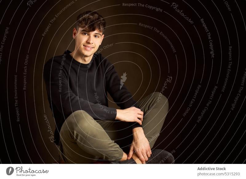 Smiling teen boy sitting on stool with crossed legs and looking at camera man smile studio appearance positive style outfit cheerful apparel teenage cloth