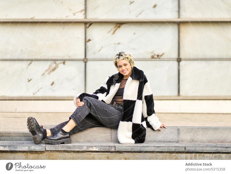 Cheerful woman sitting on stairs street style model appearance embracing knee rest city positive trendy outfit coat female young smile floor urban building fur