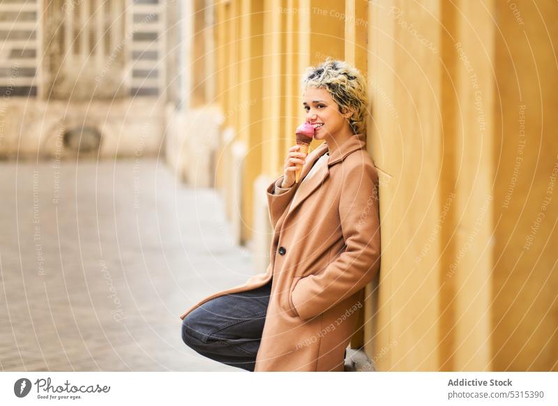 Cheerful woman eating ice cream on street smile city delicious dessert happy positive coat young urban town yummy female cone tasty building cheerful joy sweet