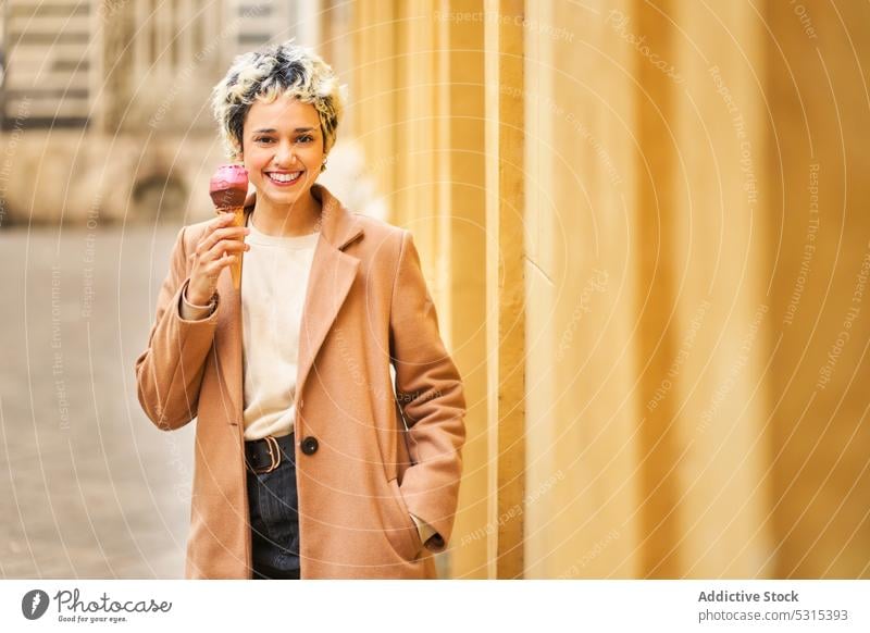 Cheerful woman eating ice cream on street smile city delicious dessert happy positive coat young urban town yummy female cone tasty building cheerful joy sweet