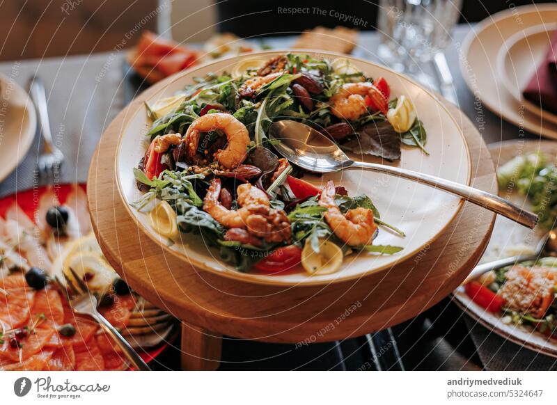 Close up festive table with fresh salad with grill seafood, greens and tomatoes on plate on wooden stand. Banquet table in restaurant with snacks, cheese appetisers and salami, holidays catering